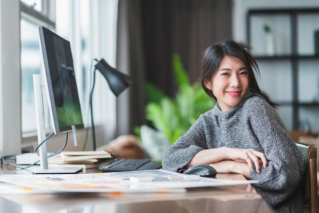 Atractiva mujer asiática inteligente mira la cámara sonriendo con confianza y alegría trabajando desde casa Una mujer asiática adulta joven trabaja con pc y un documento en papel con felicidad y disfruta del estilo de vida en casa