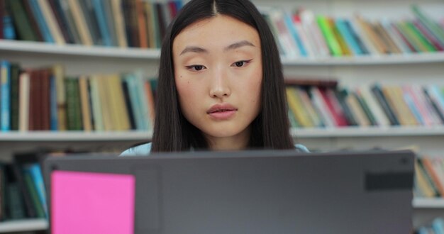 Atractiva mujer asiática leyendo un libro y escribiendo en la computadora Estudiante universitario trabaja en un ensayo en la biblioteca
