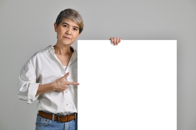 Atractiva mujer asiática de mediana edad señalando con el dedo al letrero en blanco aislado sobre fondo blanco. mirando a la camara