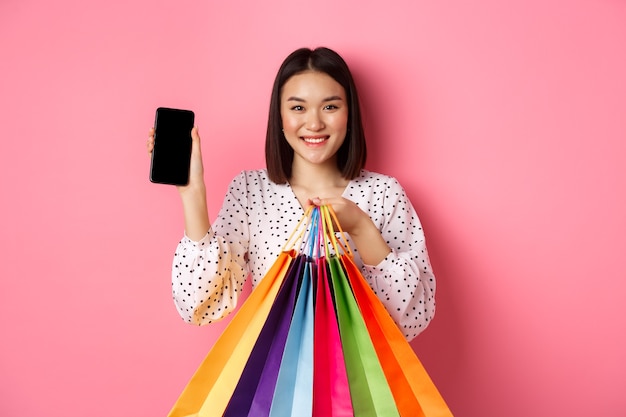 Atractiva mujer asiática que muestra la aplicación del teléfono inteligente y bolsas de compras, comprando en línea a través de la aplicación, de pie sobre rosa