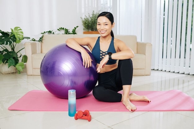 Atractiva mujer asiática sonriente sentada en la estera de yoga, apoyado en la pelota de fitness y sonriendo a la cámara