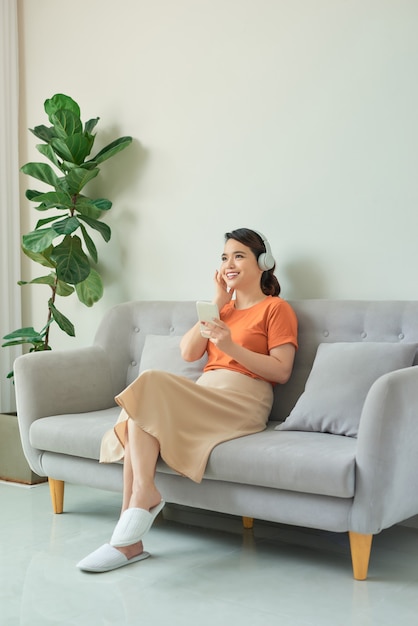 Atractiva mujer asiática usando audífonos, usando un teléfono móvil y sentado en el sofá