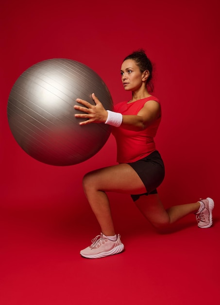 Atractiva mujer atlética haciendo estocadas hacia adelante mientras hace ejercicio con una pelota en forma sosteniéndola al nivel del pecho aislada sobre fondo rojo con espacio publicitario de copia