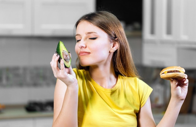 Atractiva mujer de cabello largo que duda entre aguacate fresco y hamburguesa poco saludable decide tener el concepto de fruta verde nutritiva de elegir