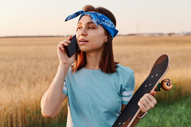 Atractiva mujer de cabello oscuro hablando por teléfono inteligente con patineta al aire libre en el campo, esperando amigos para patinar juntos, vistiendo camiseta y banda para el cabello.