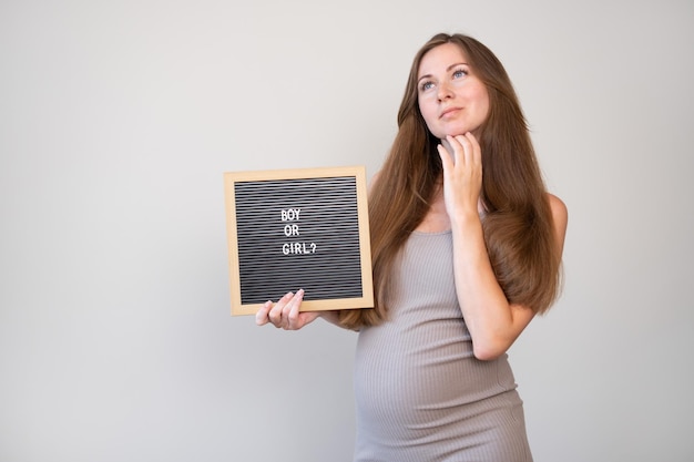 Atractiva mujer caucásica embarazada soñando pensativa sosteniendo una tabla con un niño o una niña pregunta sobre fondo gris claro Concepto de revelación de géneroCopiar espacio para texto