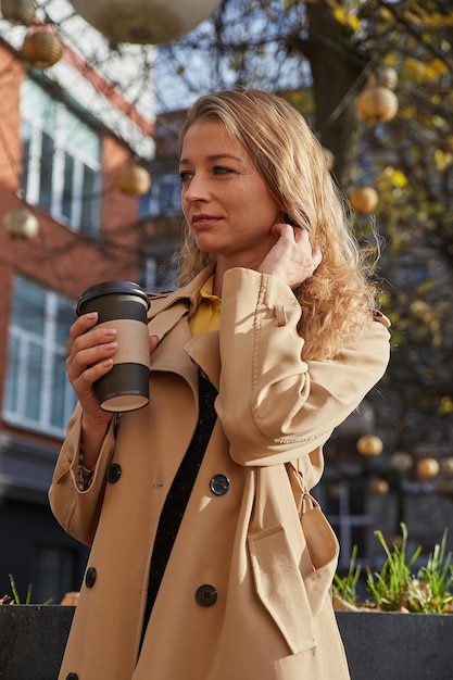 Atractiva mujer caucásica con gabardina beige sosteniendo una taza de café al aire libre