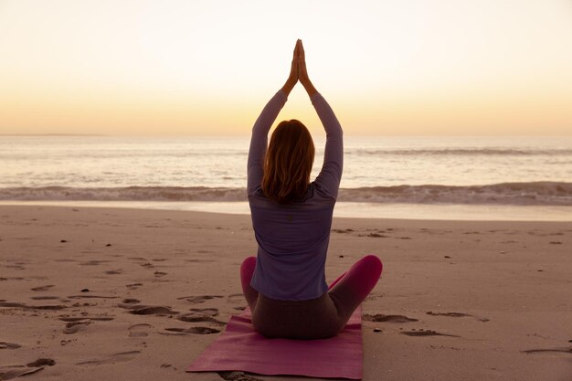 Atractiva mujer caucásica rubia disfrutando del tiempo en la playa al atardecer, practicando yoga con las manos en posición de oración, con el cielo al atardecer y el mar al fondo. Vacaciones de verano en la playa tropical.