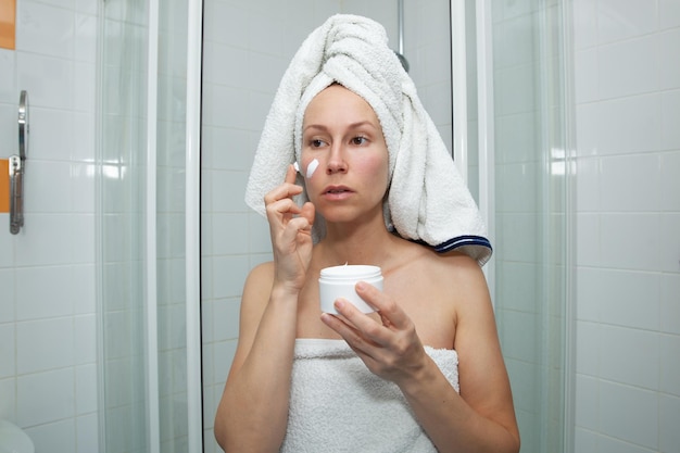 Atractiva mujer caucásica con una toalla blanca en la cabeza aplica una crema blanca en la cara después de tomar una ducha o un baño Tratamientos de spa en casa