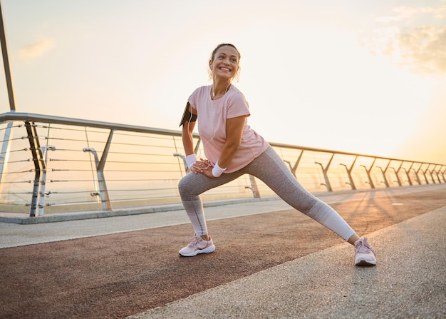 Atractiva mujer deportiva con cuerpo sano haciendo estocadas laterales, estirando los músculos de las piernas mientras hace ejercicio al aire libre en un puente de la ciudad al amanecer. Concepto de deporte, fitness, cuidado corporal y estilo de vida activo