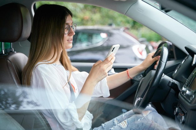 Atractiva mujer elegante con anteojos sentada en el auto y mirando la pantalla del teléfono inteligente