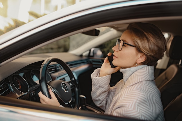 Atractiva mujer elegante sentada en el coche vestida con estilo de abrigo de invierno y gafas con teléfono inteligente