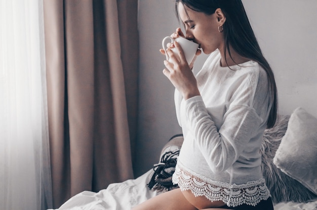 Atractiva mujer embarazada bebe té en la cama. Beber té mirando por una ventana en casa. Últimos meses de embarazo.