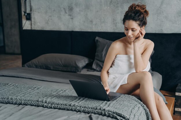 Atractiva mujer europea feliz envuelta en una toalla después de bañarse está charlando en la pc en el dormitorio