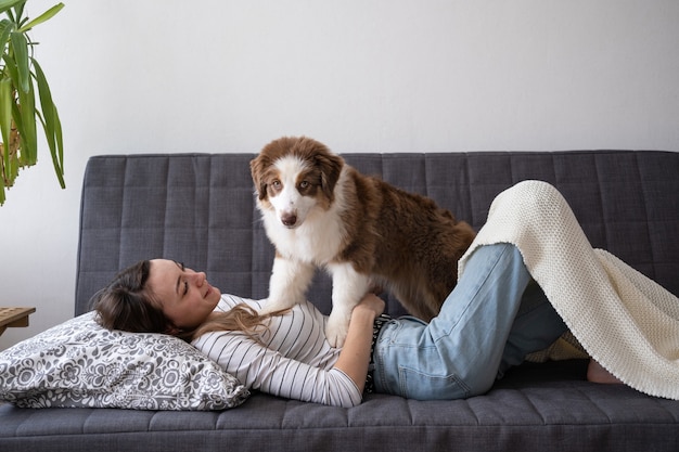 Atractiva mujer feliz con hermoso pequeño pastor australiano lindo rojo tres colores cachorro de perro acostado en el sofá. Amor y amistad entre humanos y animales.