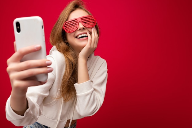 Atractiva mujer feliz sonriente joven encantadora sosteniendo y usando teléfono móvil tomando selfie vistiendo ropa elegante aislada sobre fondo de pared.