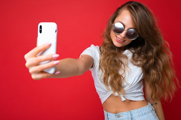 Atractiva mujer feliz sonriente joven encantadora sosteniendo y usando teléfono móvil tomando selfie