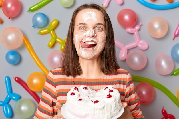 Atractiva mujer hambrienta de cumpleaños con vestido a rayas parada contra una pared gris decorada con globos coloridos sosteniendo crema para lamer pasteles de su cara mostrando la lengua afuera