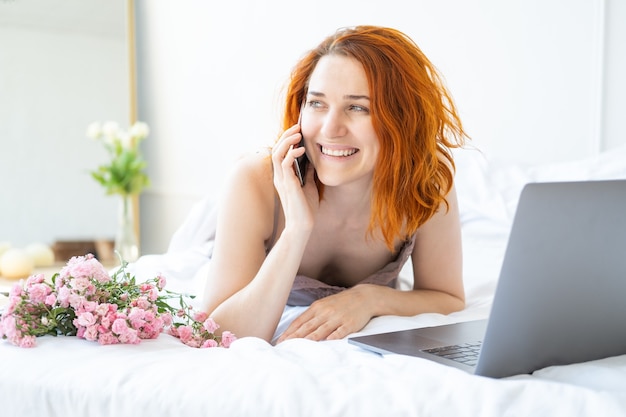 Atractiva mujer de jengibre de mediana edad en pijama acostado en la cama con teléfono y portátil