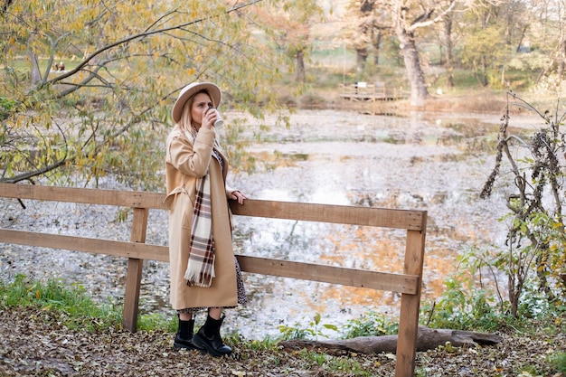 Atractiva mujer joven con abrigo beige y sombrero bebe café junto al lago en el parque de otoño en otoño