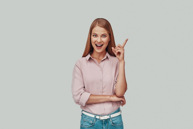 Atractiva mujer joven apuntando copia espacio y sonriendo mientras está de pie contra el fondo gris