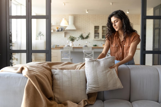 Atractiva mujer joven arreglando almohadas y tela escocesa en un cómodo sofá haciendo que su hogar sea acogedor y vacío