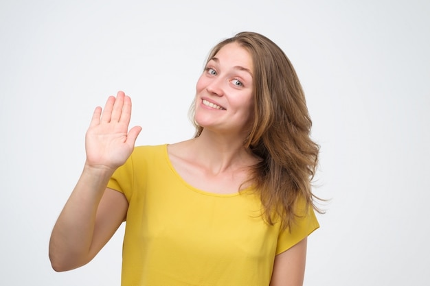 Atractiva mujer joven de aspecto amable sonriendo felizmente, diciendo Hola, Hola o Adiós, agitando la mano