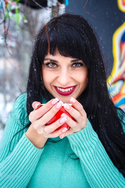 Atractiva mujer joven bebe café en la cafetería de la calle en un día nevado de invierno sonriendo mirando a la cámara