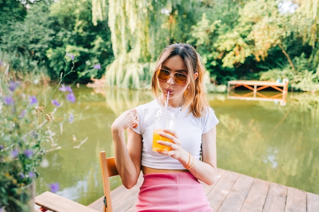 Atractiva mujer joven bebiendo limonada de naranja, descansando sobre un muelle en el lago al aire libre.