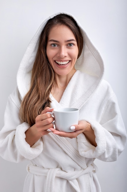 Atractiva mujer joven bebiendo té o café en la mañana vistiendo bata de baño posando sonriendo aislado ...