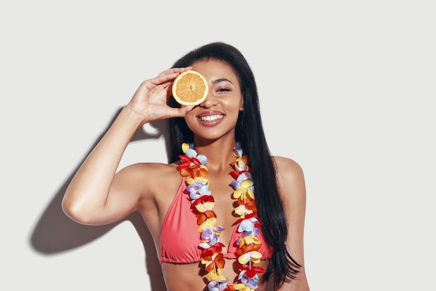 Atractiva mujer joven en bikini cubriendo los ojos con una rodaja de naranja y sonriendo mientras está de pie contra el fondo gris