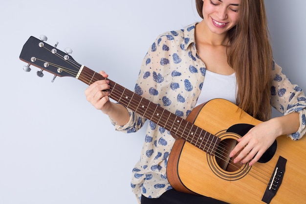 Atractiva mujer joven en blusa beige tocando la guitarra y sonriendo sobre fondo gris en estudio