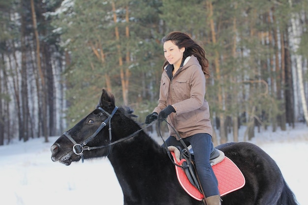 Atractiva mujer joven cabalgando sobre un caballo negro en el bosque nevado, disparo de teleobjetivo