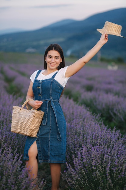 Atractiva mujer joven caminando en el campo de lavanda de verano Modelo vestida con vestido de mezclilla con sombrero de paja y bolsa