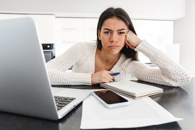 Atractiva mujer joven cansada que trabaja con la computadora portátil y documentos mientras está sentado en la cocina