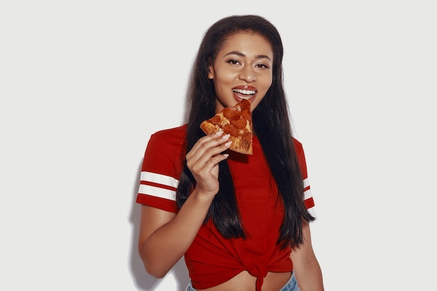 Atractiva mujer joven comiendo pizza y mirando a la cámara mientras está de pie contra el fondo gris