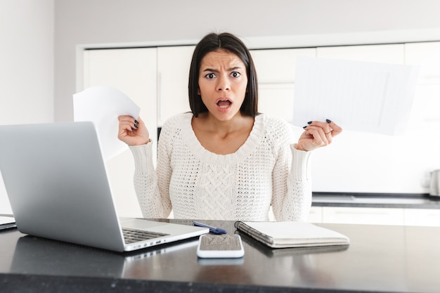 Atractiva mujer joven enojada que trabaja con la computadora portátil y documentos mientras está sentado en la cocina, gritando