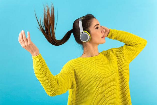 Atractiva mujer joven con estilo escuchando música en auriculares inalámbricos feliz usando suéter de punto amarillo estilo colorido moda posando aislado sobre fondo azul agitando la cola de pelo largo