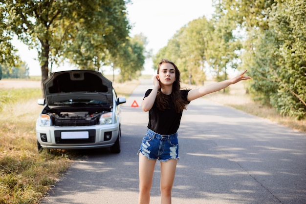 Atractiva mujer joven estudiante intenta detener el auto de los transeúntes y pedir ayuda porque su auto se ha roto