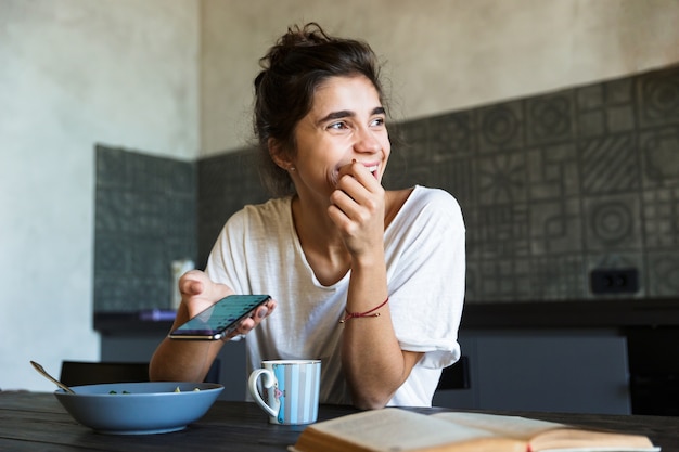 Foto atractiva mujer joven feliz con un desayuno saludable en la cocina de su casa, mensajería en el teléfono móvil