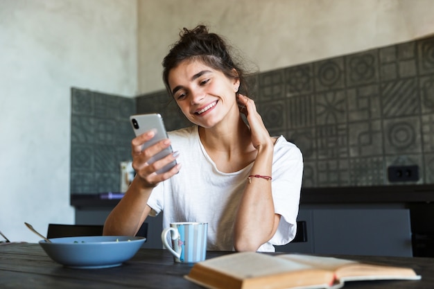 Atractiva mujer joven feliz con un desayuno saludable en la cocina de su casa, mensajería en el teléfono móvil