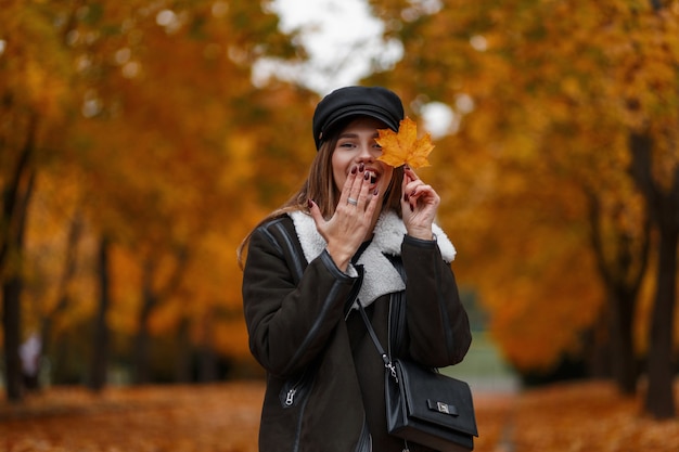 Atractiva mujer joven feliz en ropa de moda sostiene una hoja de arce amarilla cerca de la cara y cubre la boca con la mano. Hermosa chica divertida camina en el parque de otoño sobre un fondo de follaje dorado.