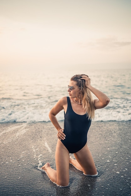 Atractiva mujer joven con gafas de sol se arrodilla en el mar a la luz del atardecer en un traje de baño negro Enfoque selectivo