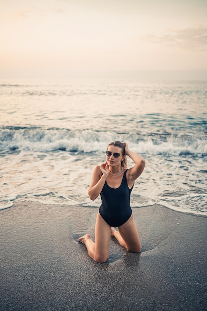 Atractiva mujer joven con gafas de sol se arrodilla en el mar a la luz del atardecer en un traje de baño negro Enfoque selectivo