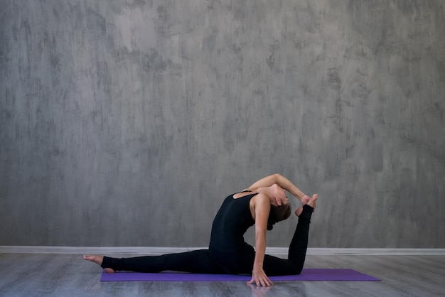 Atractiva mujer joven haciendo yoga estirando yoga en línea en casa contra gris