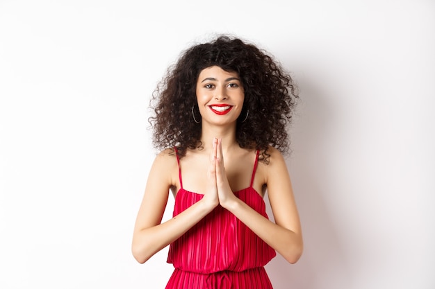 Atractiva mujer joven con labios rojos, vestido de noche, dando las gracias y sonriendo, agradecida con gesto de namaste, de pie sobre fondo blanco.