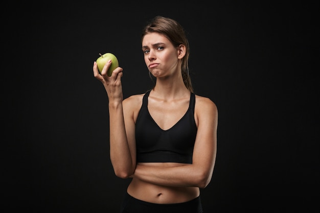 Atractiva mujer joven molesta fitness saludable vistiendo sujetador deportivo y pantalones cortos aislados sobre fondo negro, sosteniendo manzana verde