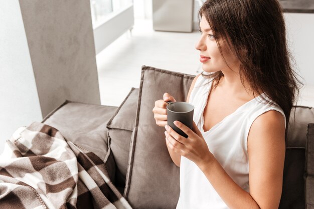 Atractiva mujer joven pacífica sentada en el sofá y tomando café