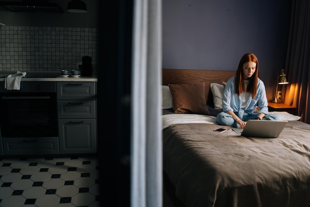 Atractiva mujer joven pelirroja haciendo videollamadas en la cámara web sentada en la cama en el lindo dormitorio. Señora feliz sonriendo en pose de loto usando computadora portátil. Bonita mujer teniendo video chat en línea con amigos