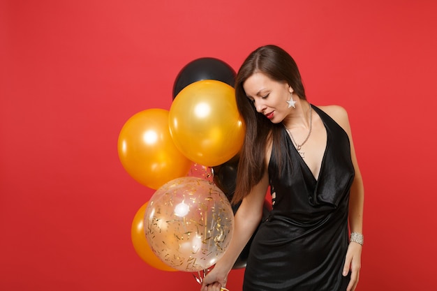 Atractiva mujer joven en un pequeño vestido negro celebrando, mirando hacia abajo sosteniendo globos de aire aislados sobre fondo rojo. Día internacional de la mujer, concepto de fiesta de vacaciones de maqueta de cumpleaños de feliz año nuevo.